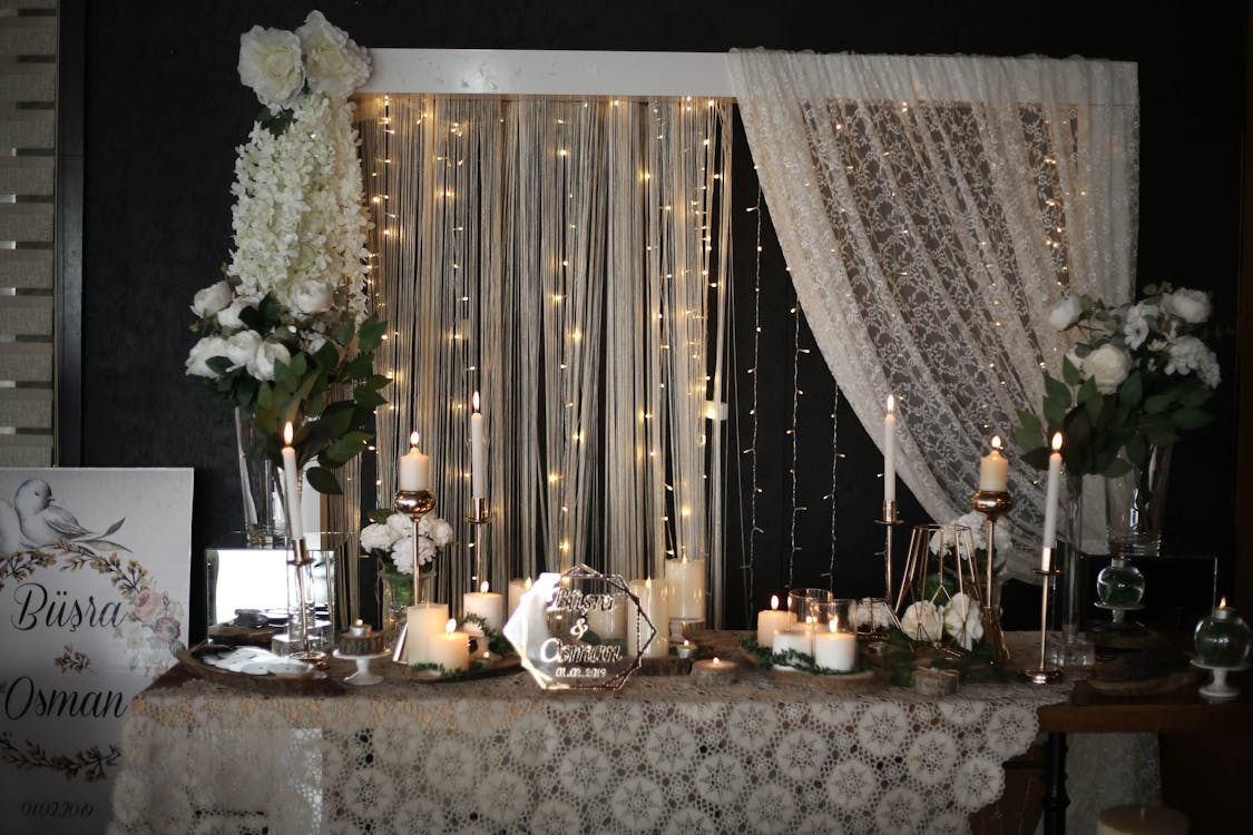 Elegant wedding table setup with floral arrangements, candles, and string light backdrop behind lace curtains